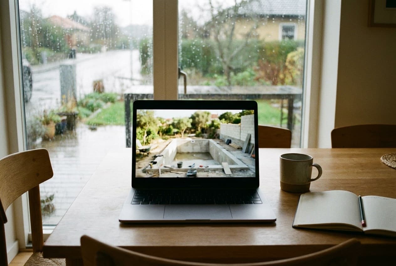Computador portátil numa mesa junto a uma janela com chuva no estrangeiro, a mostrar uma videochamada de uma piscina em construção com paredes de blocos em Portugal
